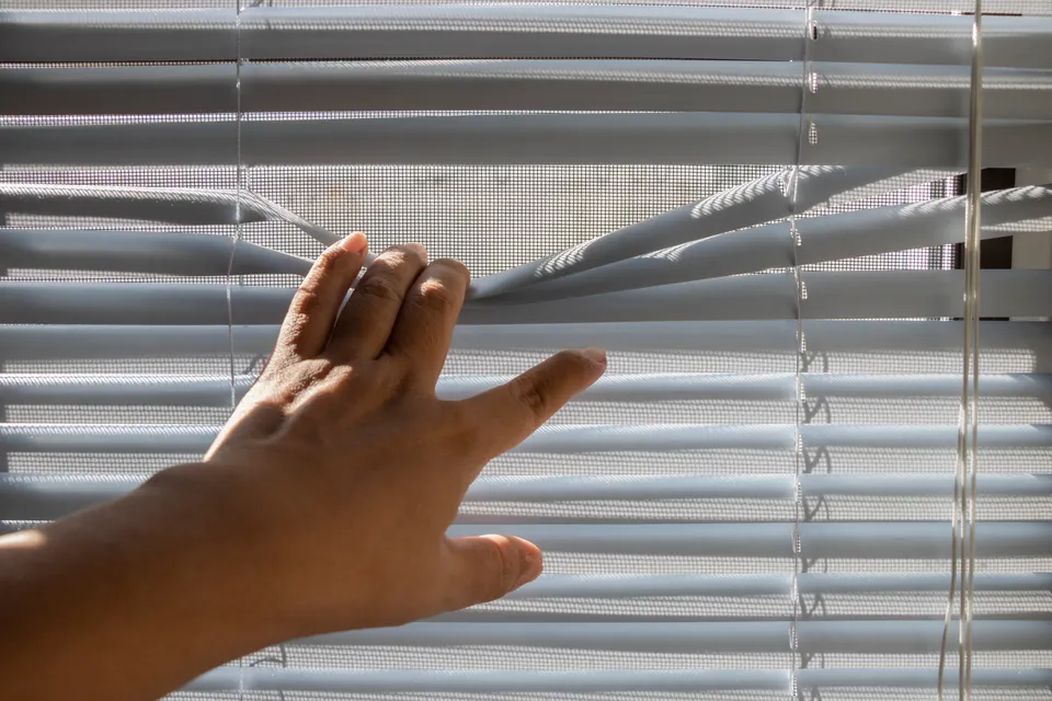 A hand adjusting pale interior window blinds.