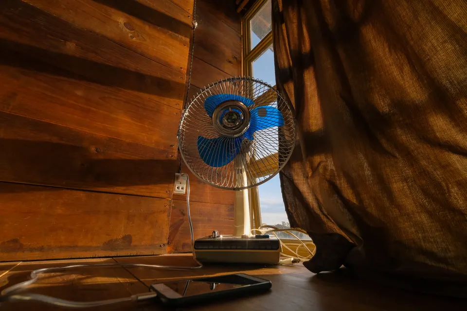 A small electric fan beside a sunlit curtain.